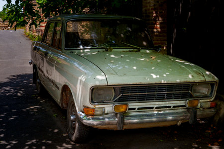Old pale green Soviet-era car standing unused in shadowy lane under overhanging tree branchesの写真素材