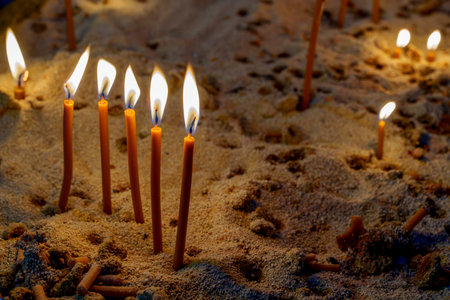 Slender wax candles stand glowing in a sand-filled tray, their warm flames flickering softlyの写真素材