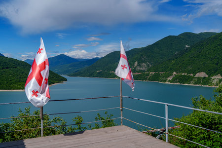 Two Georgian flags waving from wooden viewing deck overlooking blue mountain reservoir and forested hillsの写真素材