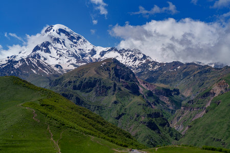 Iconic Mount Kazbek in Georgia rises above lush green foothills under bright blue sky.