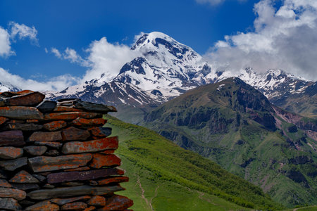Traditional stone wall in the foreground frames snowy Mount Kazbek rising above lush green hills.