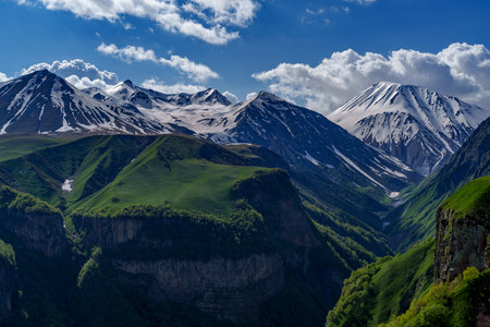 Snow covered Caucasus peaks tower above sheer green cliffs dropping into a shadowed forested gorge.
