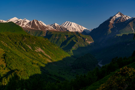 Bright morning sun illuminates snowy Caucasus peaks while a shadowed green valley stretches away...