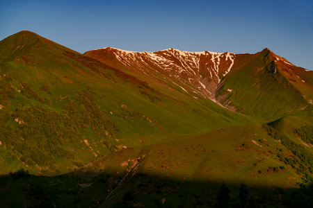 Warm sunset light paints a snowy ridgeline orange above rolling green hills and forested slopes.