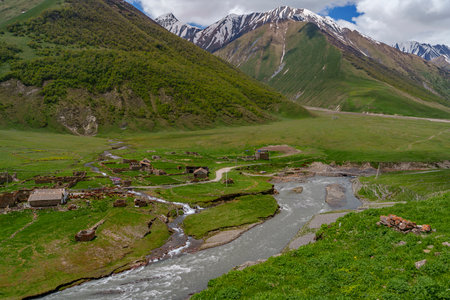 A foaming river cuts past ruined cottages and green slopes beneath rugged, snow-streaked mountain...