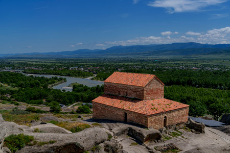 Brick church with red roof stands on rocky plateau overlooking green Mtkvari valley and mountains.
