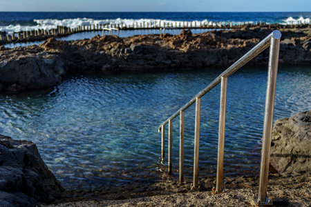 A metal railing leads into clear water, framed by dark rocks and distant surf.の写真素材