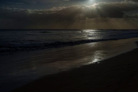 Sunlight pierces heavy clouds, forming bright reflections on wet sand and rolling surf.の写真素材