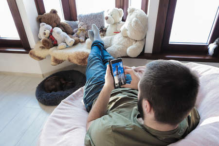 Young man, sits in comfy chair with his stretched legs lean at windowsill, is using mobile. Early morning time.の写真素材