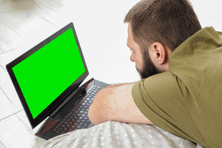 young man with beard is lying on his belly in bed and using laptop, selective focusの写真素材
