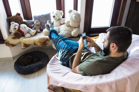 Young man, sits in comfy chair with his stretched legs lean at windowsill, is using mobile. Early morning time.の写真素材