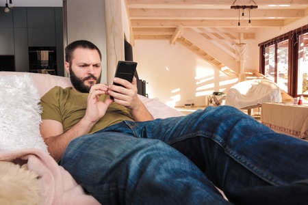 Young man, sits in comfy chair with his stretched legs, is using mobile. Early morning time.の写真素材