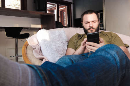 Young man, sits in comfy chair with his stretched legs, is using mobile. Early morning time.の写真素材