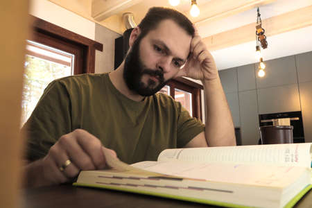 Young man with beard sitting in kitchen and reading a book. selective focus. Close up shot.の写真素材