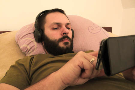 Young man with beard lie in bed and listening music on headphones while using his mobile. selective focus .. Daytime.の写真素材