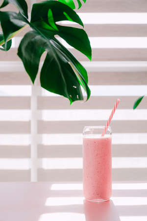 Pink smoothie in a glass with a straw on the background of a palm leaf.の写真素材
