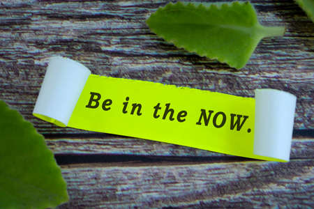 Be in the now words on yellow note with leave on wooden desk.の写真素材