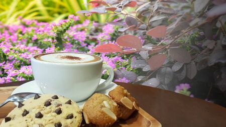 Hot cup of coffee in a tray with a dessert on a wooden table with a flower garden backdropの写真素材