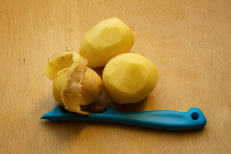 Three potato lying on an old wooden table, two of them are peeled and the third is chosen.partly. Next is blue potato peeler, with a piece of potato skin.Horizontal view.の写真素材