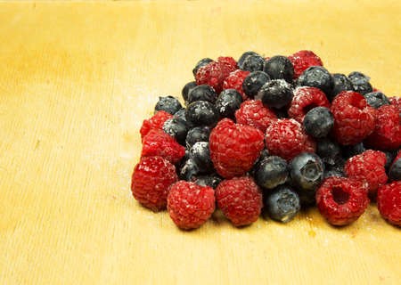 On the old wooden board lies a handful of frosted highbush blueberries and raspberries. Close horizontal view.の写真素材