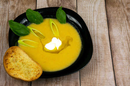Photo of cream of leek soup with fresh pieces of leek,basil leaves,sour cream and toast bread with garlic butter.Soup in black plate on wooden background.Horizontal view from top.Closeup.の写真素材