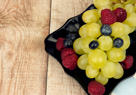 A photo close up grapes, raspberries and blueberries on a black saucer standing on a wooden table. Copy space on the left side.Flat horizontal view from above.の写真素材