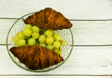 Bunch of green grapes and two croissants in a glass bowl on a wooden old table style vintage.Flat horizontal view from above.の写真素材