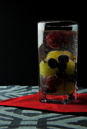 Sparkling water with raspberries, blueberries and grapes in a high glass on red serwetve and black background. Clearly visible gas bubbles on the fruit. Close, vertical view.の写真素材