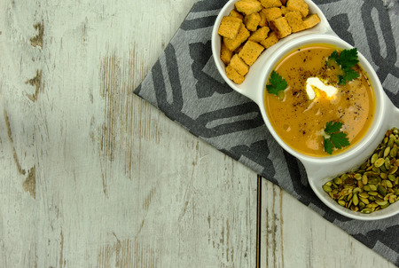 White dish with pumpkin soup, pumpkin seeds and small, baked croutons of bread, prepared the fresh leaves of parsley and cream and pepper on old wooden table., copy space on the left side.Flat horizontal view from above.の写真素材