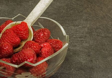 Glass bowl with fresh fruit raspberries and old wooden spoon on stone, dark table. Cloes horizontal...