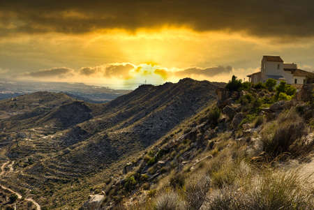 Sunset over the mountains and the house, on top of the mountain, illuminated by the orange light of the hiding sun.Spain, Alicante province.Horizontal view.の写真素材