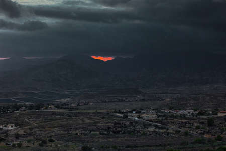 Heavy storm clouds at dusk over the mountains. A piece of red sun disc can be seen through the...