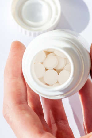 Man looking into a small open pill container with white pills inside in a sterile environment, holding it in his hand, examining it. Taking medication, drug addiction, supplementation simple conceptの写真素材