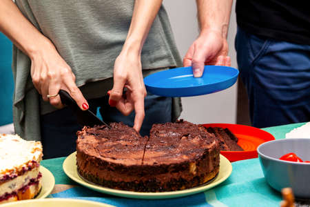 Simple sweet brown chocolate cake being cut into pieces with a knife by a woman man waiting with empty blue plate Treating someone with cake birthday party concept splitting slicing a cake anonymousの写真素材