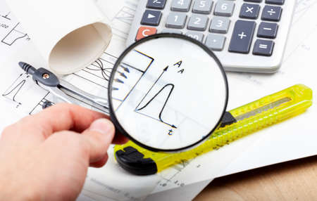 Scientist, architect or electrician holding a magnifying glass and inspecting analyzing data on a graph, chart. Engineering abstract concept, notes, blueprints and various tools in the backgroundの写真素材