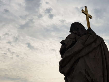 A statue of a saint holding a metallic cross seen from the back with bright sky full of clouds as a background. Christianity conceptの写真素材