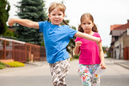 Two happy little children, sisters, siblings walking, girls jumping happily towards the camera with arms spread wide. Kids family walk, town area, natural lifestyle scene, simple portrait outdoorsの写真素材
