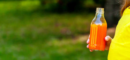 Anonymous little child, girl holding a glass bottle of healthy orange carrot vegetable juice, banner, hand closeup, copy space on the left. Nutritious drinks full of vitamins, healthy kids, beveragesの写真素材