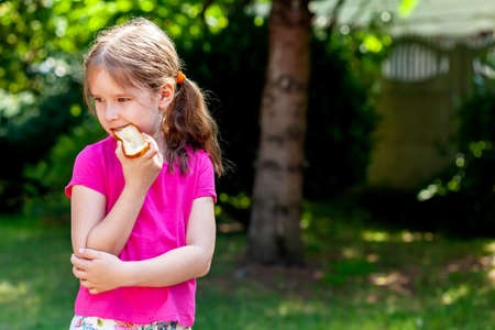 Little school age girl, young child eating, biting a single slice of bread with butter, copy space, outdoors portrait, lifestyle. Children and nutrition, food, eating habits, wheat products conceptの写真素材
