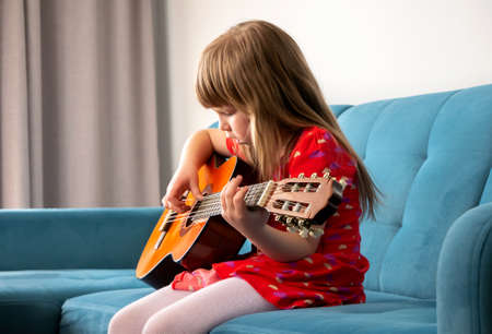 Little girl, young school age child playing an acoustic guitar, practicing a musical instrument sitting on a sofa at home, interior, side view. Musical education, hobby, childs talent abstract conceptの写真素材