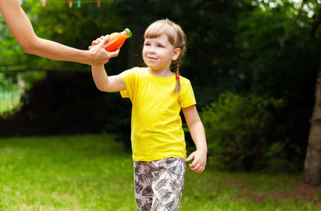 Mother giving her little daughter a glass bottle full of healthy carrot juice, garden, outdoors. Children nutrition, nutritious vegetable juice full of vitamins, parental care concept, lifestyle shot