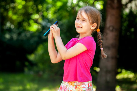 Cheerful school age child, happy little young girl holding her smartphone, mobile phone in hands smiling, outdoors portrait, copy space, closeup. Children and technology, telecommunication, lifestyleの写真素材