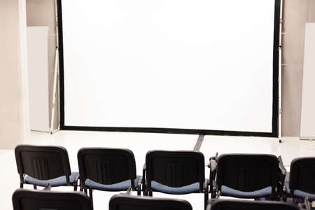 Rows of office chairs in front of a blank corporate multimedia presentation screen, vacant conference room interior, empty auditorium, public event, employee training, education, nobody, copy space.の写真素材