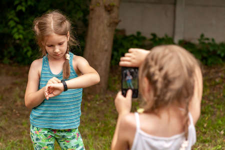 Two young little children, school age kids using electronic gadgets, kids and technology outdoors shot. Smart fit band and a smartphone, taking photos with a mobile phone concept, people, lifestyleの写真素材