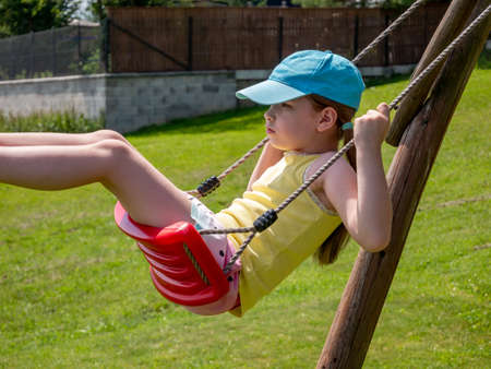 Little caucasian girl, young school age child sitting on a swing, side view, closeup, portrait. Having fun outdoors alone, playing outside by herself. Childhood leisure and relaxation simple conceptの写真素材
