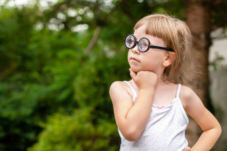 Young clever intelligent school age child, little girl in big quirky glasses thinking, wondering, deep in thought, copy space, outdoor portrait, face closeup. Hand under chin, one person, educationの写真素材