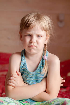 One single offended child, young little school age girl sitting on the bed at home with arms crossed, unhappy rebellious kid simple vertical portrait, face closeup, frown, dissatisfied expressionの写真素材
