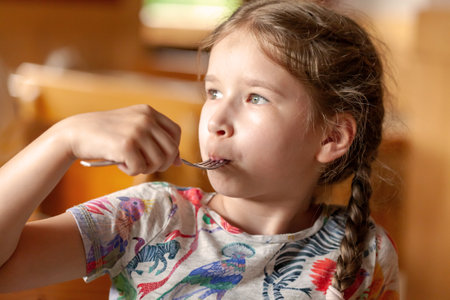 Young school age girl, little child eating her meal, holding a fork in mouth, indoors portrait, face closeup, shallow depth of field. Children, childhood nutrition, appetite, healthy lifestyle conceptの写真素材