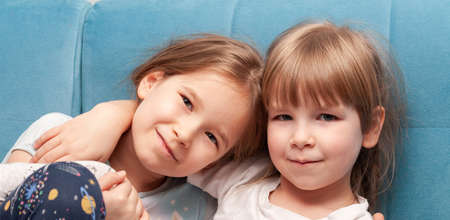 Two young cheerful elementary primary school age girls smiling, sisters, siblings hugging, sitting together on a sofa, portrait, face closeup. Family happiness, togetherness and bonding simple conceptの写真素材