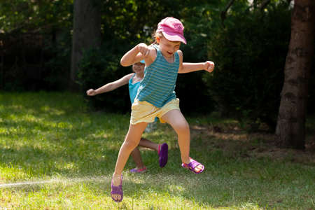 Two active joyful children, girls playing with water in the backyard jumping, running around, sisters siblings summer leisure, water hose, having fun outdoors, leisure, recreation, togethernessの写真素材